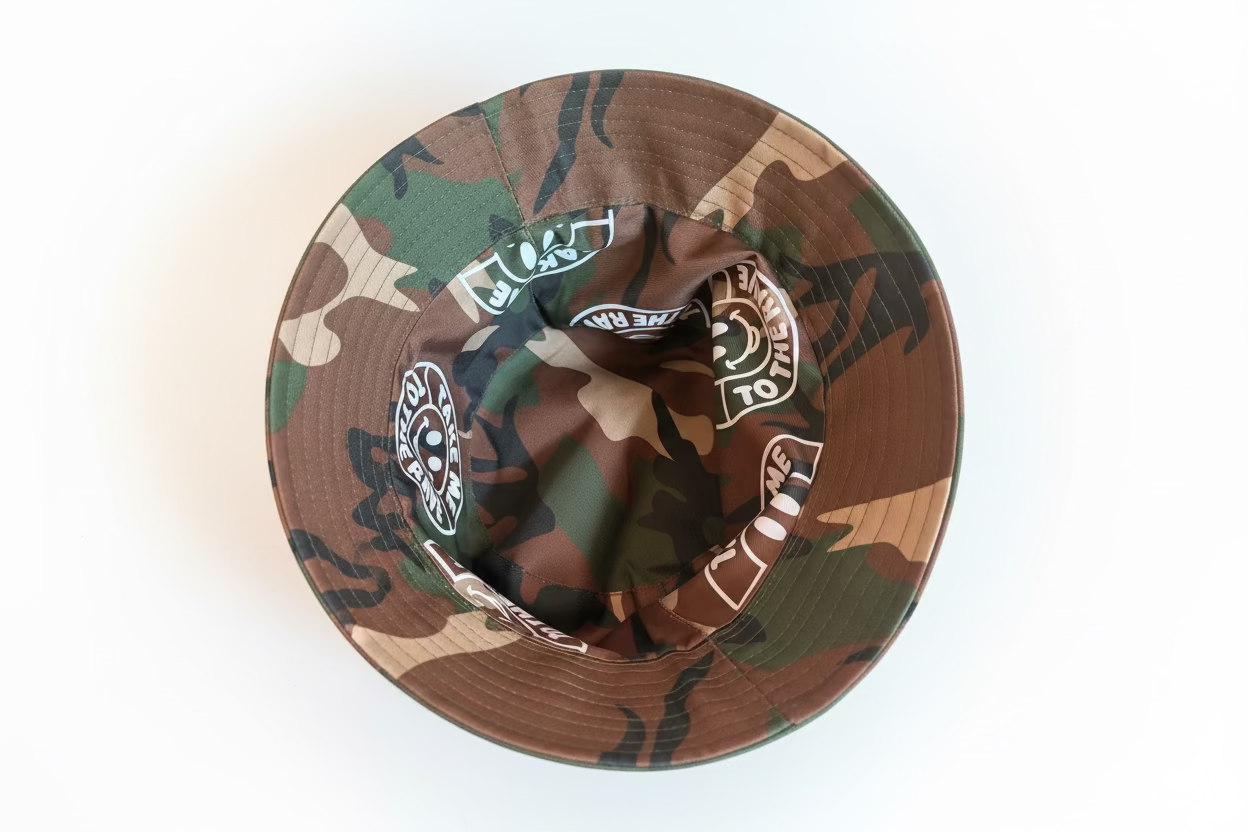 Close-up of the inside of a camouflage bucket hat, showing a brown, green, and tan camo pattern with white printed circular logos around the inner brim.