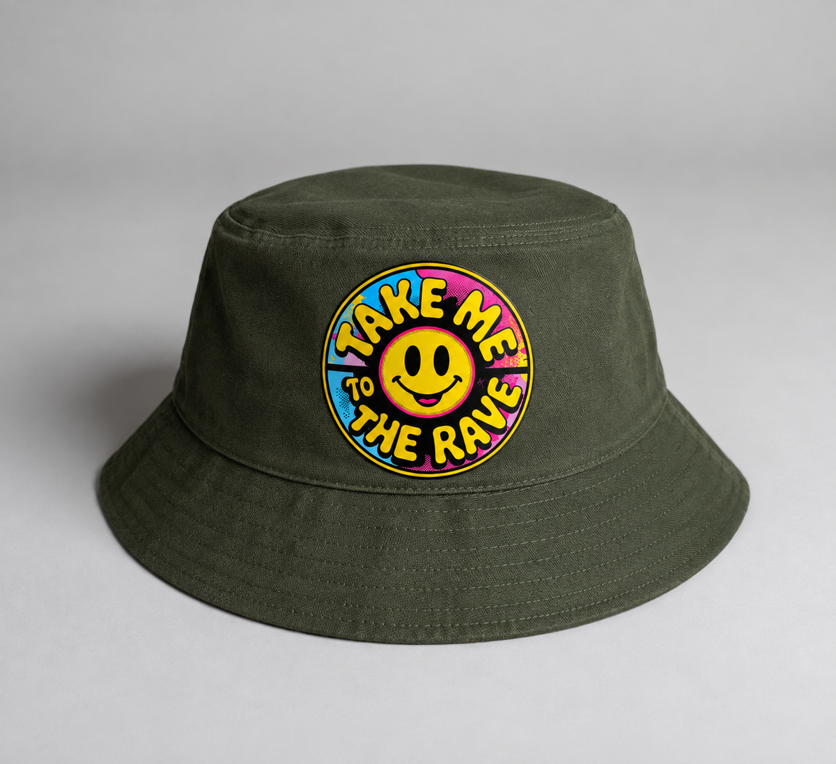 An olive green bucket hat with a circular embroidered patch featuring a yellow smiley face and the text 'TAKE ME TO THE RAVE' in neon colors, set against a neutral gray background.
