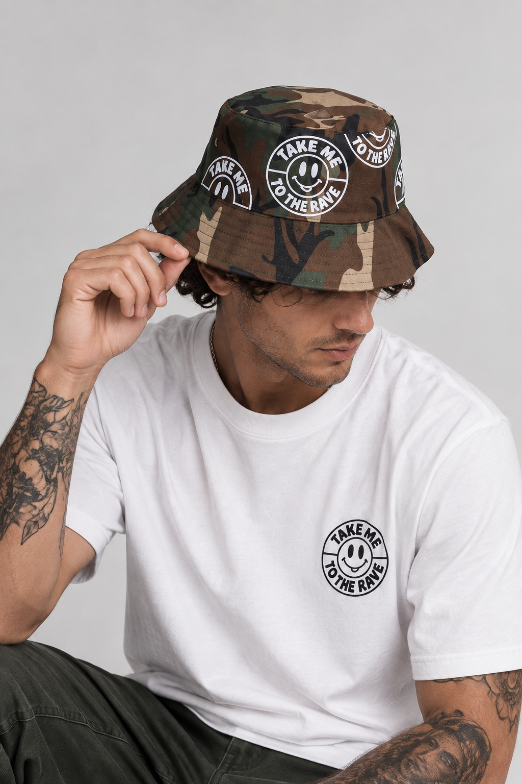 A man wearing a camouflage take me to the rave bucket hat with white smiley face graphics and text, paired with a white t-shirt, posed against a light gray studio background.